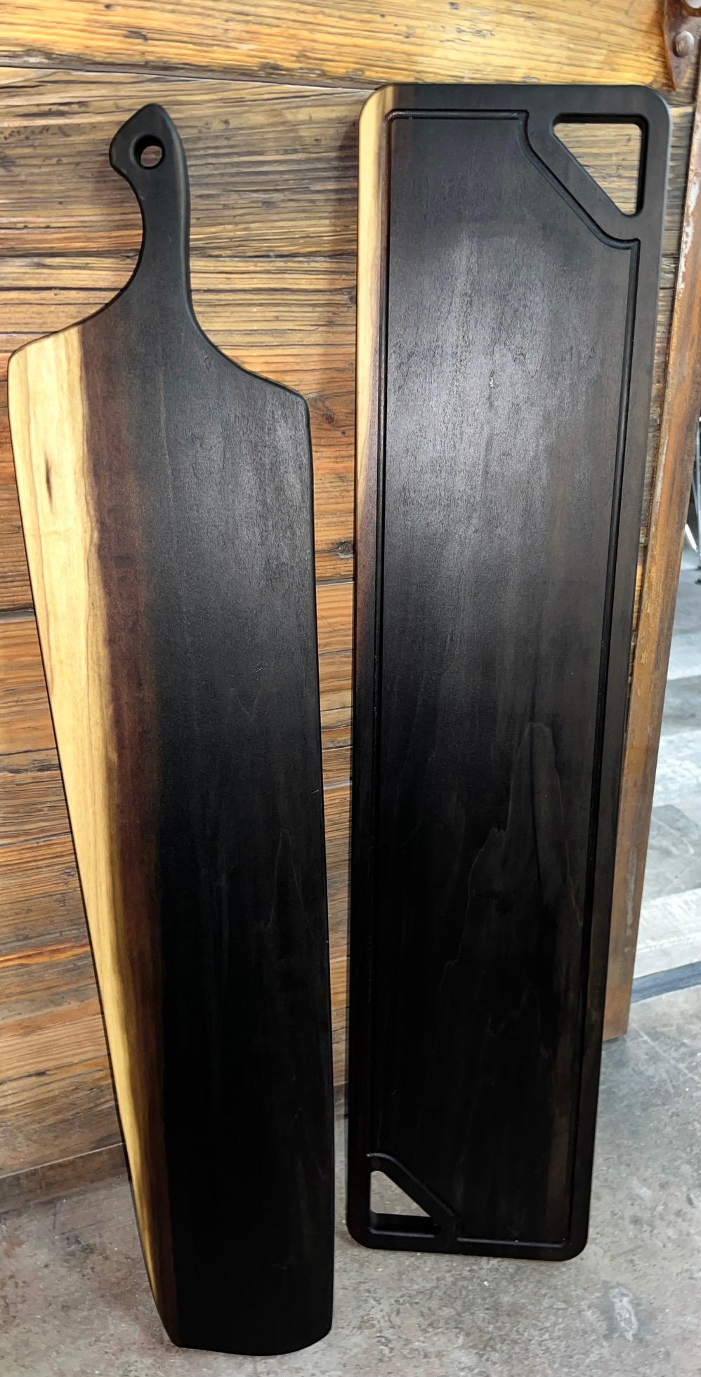 Dos tablas de madera ebonizada recargadas sobre un muro: una con mango de orificio, otra rectangular con esquina biselada.